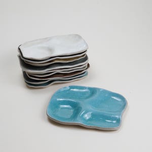 Könnte beinhalten: Ein Stapel unregelmäßig geformter Keramikplatten in verschiedenen Farben, darunter Weiß, Beige, Grau und Braun. Vor dem Stapel liegt eine einzelne türkisfarbene Platte mit drei Vertiefungen. Die Platten haben eine glatte, glänzende Oberfläche.