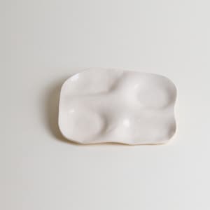 Könnte beinhalten: Eine weiße Keramikschale mit einer organischen, wellenförmigen Form. Die Schale hat eine glatte, glänzende Oberfläche und weist drei leicht erhöhte Bereiche auf, die eine einzigartige, strukturierte Oberfläche schaffen. Das Gesamtdesign ist minimalistisch und modern.