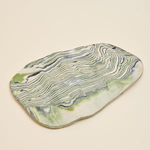 May include: An oval-shaped ceramic tray featuring a marbled pattern of white, sage green, and dark navy blue. The surface displays intricate, wavy, layered lines that create a topographical effect across the entire face of the dish.
