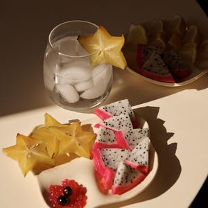 Puede incluir: Una bebida refrescante con hielo y una rodaja de carambola. Rodajas de fruta del dragón y carambola están dispuestas en un plato blanco, junto con un pequeño plato de bayas rojas. Rodajas de limón en el fondo.