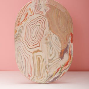 May include: An oval ceramic tray featuring a marbled pattern with swirling lines in shades of cream, soft orange, muted green, and hints of purple. The surface has a smooth, glossy finish with a raised edge, showcasing a fluid, organic design.