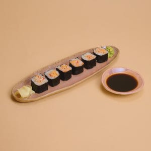 Puede incluir: Rollos de sushi con relleno naranja y envoltura de algas marinas dispuestos en un plato largo de color marrón claro. Un pequeño plato de salsa oscura y una porción de wasabi verde acompañan el sushi. Hay jengibre encurtido.