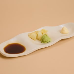 May include: A white, wavy-edged ceramic serving dish with soy sauce, wasabi, ginger, and a dollop of a creamy condiment. The dish is set against a neutral background, highlighting the colors and textures of the food.
