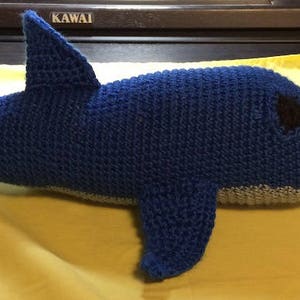 May include: A blue crocheted dolphin plush toy with a gray belly and black eyes. The toy is lying on a yellow fabric surface.