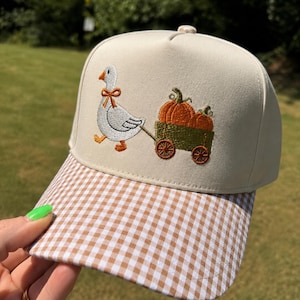 May include: A beige baseball cap featuring an embroidered design of a white goose with an orange bow around its neck, pulling a green wagon filled with orange pumpkins. The cap has a brown and white gingham patterned brim and a button on top.
