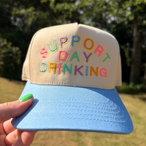 Könnte beinhalten: Eine beige Baseballkappe mit hellblauem Schirm. Auf der Vorderseite befindet sich der gestickte Schriftzug "SUPPORT DAY DRINKING" in bunten Buchstaben, darunter rosa, gelbe, grüne, blaue und violette Fäden auf dem neutralen Stoff.