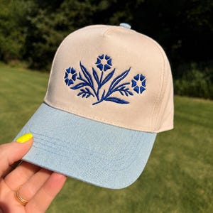 May include: A baseball cap featuring a cream-coloured crown and a light blue denim-textured brim. The front panel displays an embroidered design of three blue flowers with stems and leaves centred on the fabric. The cap has a structured front panel.