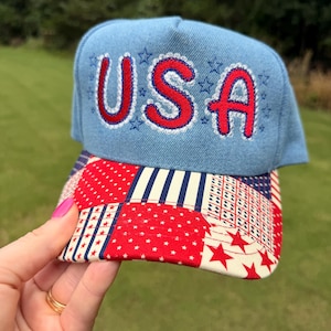 以下が含まれることがあります： 赤色の刺繍でUSAと書かれ、銀色の縁取りが施された水色の野球帽。つばの部分には、赤、白、青の布地を組み合わせたパッチワーク模様が施されており、全体に星やストライプのデザインがあしらわれています。