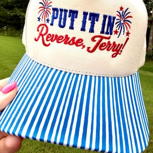 May include: A beige baseball cap with a blue and white vertical striped brim. The front features embroidered text that reads "PUT IT IN Reverse, Terry!" in blue and red, flanked by two small red and blue firework designs on each side.