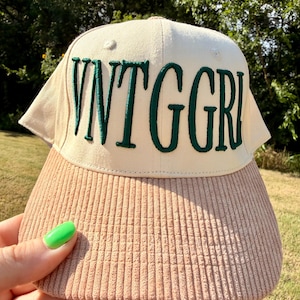 May include: A baseball cap with a cream-colored crown and a tan corduroy brim. The front of the crown features the text VNTGGR in dark green embroidered lettering. The cap has a structured design with a button on top and visible stitching.