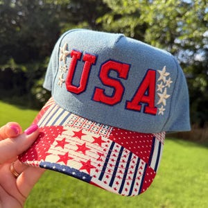 May include: A light blue denim baseball cap featuring the red embroidered text USA on the front panel, flanked by white embroidered stars. The brim displays a patchwork pattern of red, white, and blue fabric with star and stripe motifs.