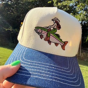 May include: A cream-coloured baseball cap with a blue denim brim featuring white contrast stitching. The front panel displays an embroidered graphic of a figure with long hair wearing a cowboy hat and boots, riding on the back of a large rainbow trout.