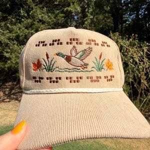 May include: A beige corduroy baseball cap featuring an embroidered mallard duck in flight over water, flanked by cattails and autumn leaves. The design includes geometric brown patterns above and below the duck, with a white braided cord trim.