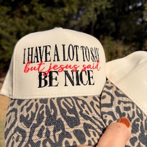 May include: A cream-colored baseball cap with a leopard print brim. The front panel features embroidered text in black and red that reads: I HAVE A LOT TO SAY but jesus said BE NICE. The cap has a structured crown and a curved visor.