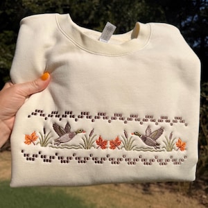 May include: A cream-colored crewneck sweatshirt featuring an embroidered horizontal band. The design depicts two mallard ducks in flight, surrounded by green reeds, cattails, and orange maple leaves, framed by rows of small brown stitched squares.