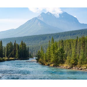 May include: A scenic landscape featuring a turquoise river winding through a lush green forest. Towering mountains with snow-capped peaks rise in the background under a clear blue sky, creating a serene natural vista.
