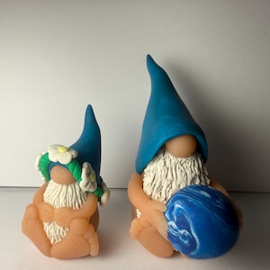 Op de afbeelding: Twee handgemaakte kabouterbeeldjes. Eén kabouter draagt een blauwe hoed en houdt een blauw-witte gemarmerde bal vast. De andere kabouter draagt een blauwe hoed en een bloemenkrans. Beide kabouters hebben witte baarden en zijn gemaakt van perzikkleurige klei.