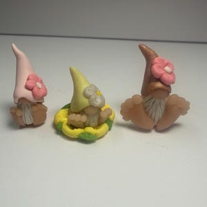 May include: Three miniature gnome figurines. One gnome wears a pink hat with a pink flower, another has a yellow hat with a white flower, and the third has a brown hat with a pink flower. Each gnome has a white beard and is sitting with their legs spread.