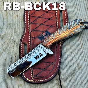 May include: A custom straight razor with a patterned handle and a stainless steel blade, resting on a brown leather sheath. The blade is engraved with "WA". The sheath is stitched with white thread and has a diamond pattern. The text "RB-BCK18" is at the top.