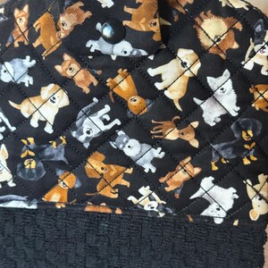 May include: A black hanging kitchen towel with a quilted top featuring a repeating pattern of various dog breeds in shades of brown, white, and gray. The bottom portion is a textured black towel. A black button secures the hanging loop.