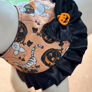 May include: A pet harness featuring a peach-colored fabric with a pattern of black pumpkins and white tiger illustrations. The harness is accented with a black satin ruffled trim and a small orange pumpkin-shaped button attached to a black bow.