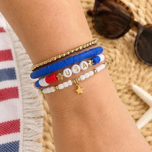Op de afbeelding: Een stapel kralenarmbanden in rood, wit, blauw en goud. Eén armband spelt "USA" met witte letterkralen. Een andere armband heeft een gouden sterbedel. Zonnebril en zeester zijn op de achtergrond te zien.