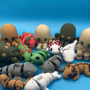 May include: A collection of 3D-printed animal figurines in various colors, including octopuses, axolotls, frogs, and dolphins. The figurines are arranged on a solid blue background, showcasing their detailed designs and playful nature.