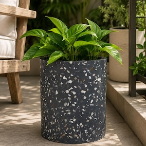 May include: A cylindrical dark gray planter with a speckled pattern, filled with vibrant green leafy plants. The planter is made of a composite material and sits on a stone patio. The overall theme is home and garden decor.