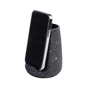 May include: A black smartphone with a silver frame is displayed in a speckled gray phone holder. The holder has a rounded base and a cylindrical opening. The phone is angled slightly, showcasing its screen.