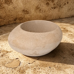 May include: A smooth, rounded stone bowl with a light beige, speckled texture. The vessel features an organic, asymmetrical shape with a wide, open top and a solid, thick-walled construction, showcasing a natural, porous surface finish throughout.