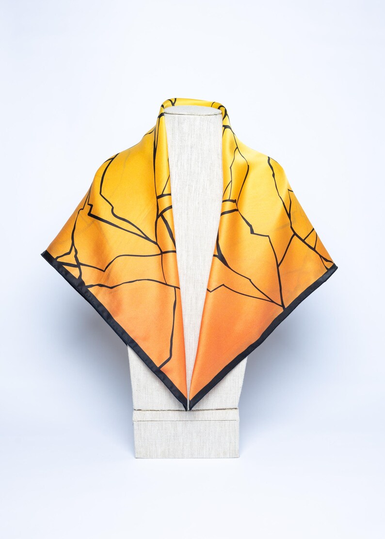 K&ouml;nnte beinhalten: Quadratisches Seidentuch mit einem Farbverlauf von Gelb zu Orange. Schwarze Linien bilden ein geometrisches Muster auf dem Tuch. Das Tuch hat einen schwarzen Rand und wird auf einem Mannequinkopf pr&auml;sentiert.
