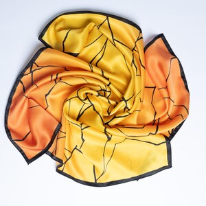 K&ouml;nnte beinhalten: Ein Seidenschal in Gelb- und Oranget&ouml;nen mit einem schwarzen geometrischen Muster und schwarzem Besatz. Der Schal ist kunstvoll drapiert und unterstreicht seine flie&szlig;ende Textur und sein Design. Ein modisches Accessoire.
