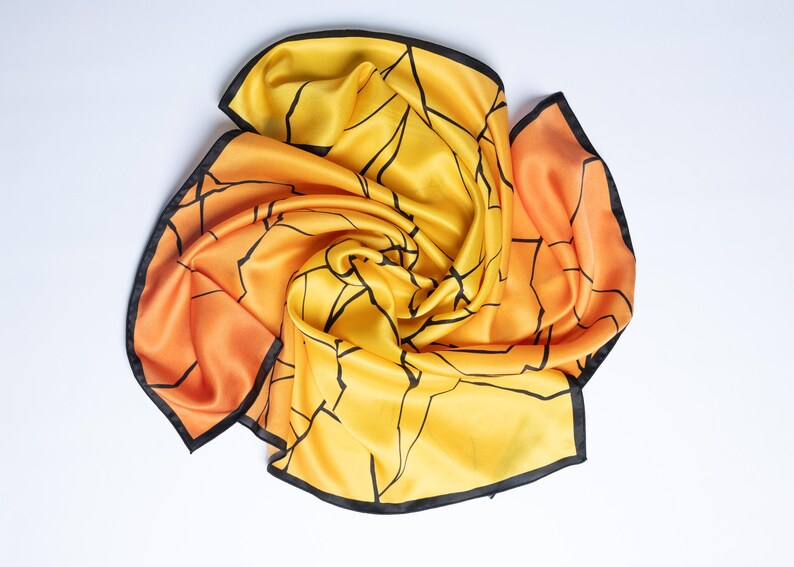 K&ouml;nnte beinhalten: Ein Seidenschal in Gelb- und Oranget&ouml;nen mit einem schwarzen geometrischen Muster und schwarzem Besatz. Der Schal ist kunstvoll drapiert und unterstreicht seine flie&szlig;ende Textur und sein Design. Ein modisches Accessoire.