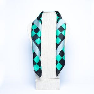 Op de afbeelding: Een zijden sjaal met een geometrisch patroon van zwarte en turquoise ruiten, tegen een lichtblauwe achtergrond. De sjaal wordt getoond op een witte paspop, waarbij het ontwerp en de drapering worden benadrukt.