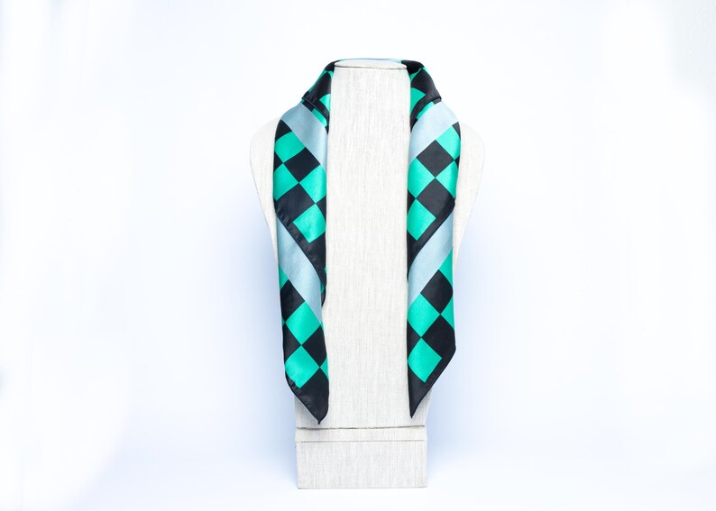 Op de afbeelding: Een zijden sjaal met een geometrisch patroon van zwarte en turquoise ruiten, tegen een lichtblauwe achtergrond. De sjaal wordt getoond op een witte paspop, waarbij het ontwerp en de drapering worden benadrukt.