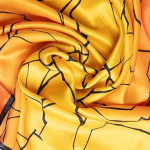 K&ouml;nnte beinhalten: Seidenschal in Gelb- und Oranget&ouml;nen, verziert mit einem geometrischen Muster aus schwarzen Linien. Der Stoff ist drapiert und zeigt seine flie&szlig;ende Textur. Die Kanten des Schals sind mit einem schwarzen Rand versehen.