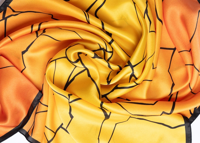 K&ouml;nnte beinhalten: Seidenschal in Gelb- und Oranget&ouml;nen, verziert mit einem geometrischen Muster aus schwarzen Linien. Der Stoff ist drapiert und zeigt seine flie&szlig;ende Textur. Die Kanten des Schals sind mit einem schwarzen Rand versehen.