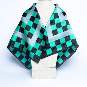 Op de afbeelding: Een vierkante sjaal met een zwart-teal groen dambordpatroon, weergegeven op een mannequin. De sjaal heeft een lichtblauwe rand en is gemaakt van een zijdeachtig materiaal. Een modeaccessoire.