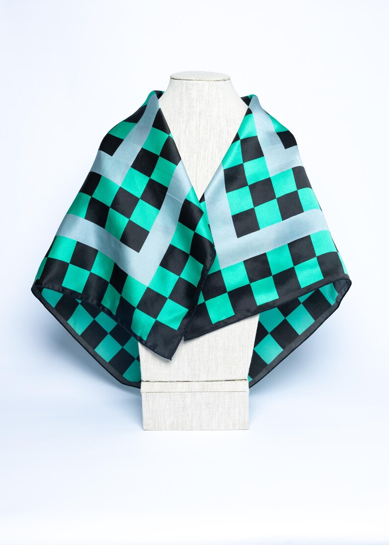 Op de afbeelding: Een vierkante sjaal met een zwart-teal groen dambordpatroon, weergegeven op een mannequin. De sjaal heeft een lichtblauwe rand en is gemaakt van een zijdeachtig materiaal. Een modeaccessoire.