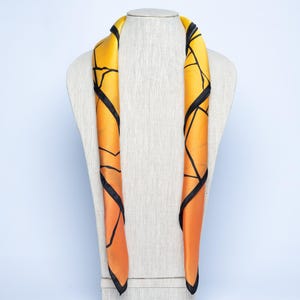 K&ouml;nnte beinhalten: Ein Seidenschal mit geometrischem Design. Der Schal hat einen Farbverlauf von Gelb zu Orange mit schwarzen Linien. Er ist &uuml;ber eine Schaufensterpuppe drapiert. Der Schal ist ein Modeaccessoire.