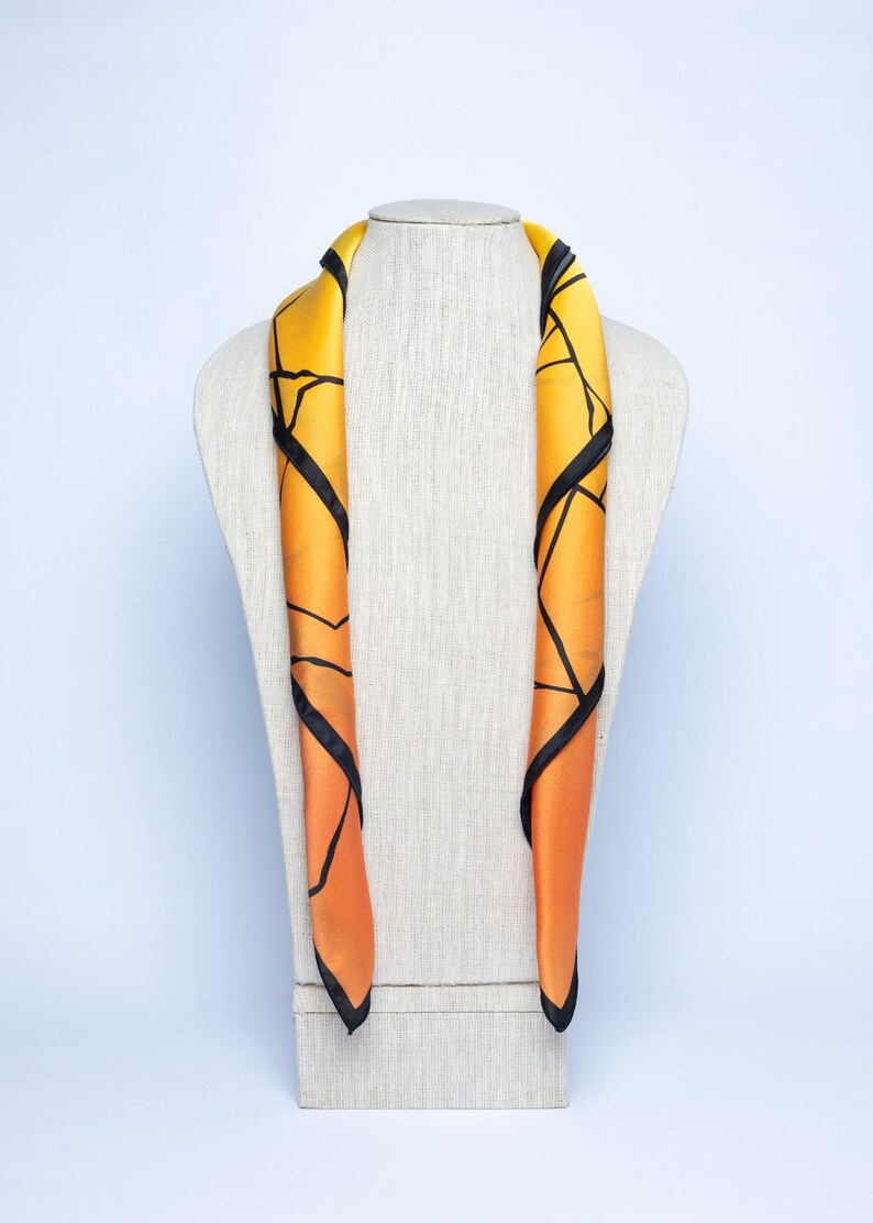 K&ouml;nnte beinhalten: Ein Seidenschal mit geometrischem Design. Der Schal hat einen Farbverlauf von Gelb zu Orange mit schwarzen Linien. Er ist &uuml;ber eine Schaufensterpuppe drapiert. Der Schal ist ein Modeaccessoire.
