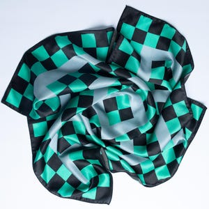 Op de afbeelding: Een vierkante sjaal met een geblokt patroon in zwart, teal en grijs. De sjaal is gemaakt van een zijdeachtig materiaal en is gedrapeerd op een witte ondergrond. Het ontwerp heeft een herhalend patroon van vierkanten.