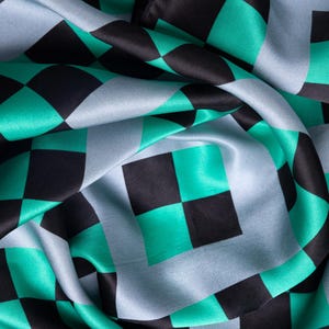 Op de afbeelding: Close-up van een zijdeachtige stof met een herhalend dambordpatroon. Het patroon heeft vierkanten in zwart, teal en lichtblauw. De stof is gedrapeerd, waardoor plooien en schaduwen ontstaan die diepte en textuur aan het ontwerp toevoegen.