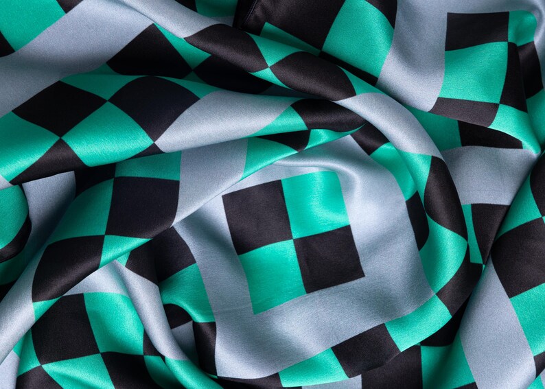 Op de afbeelding: Close-up van een zijdeachtige stof met een herhalend dambordpatroon. Het patroon heeft vierkanten in zwart, teal en lichtblauw. De stof is gedrapeerd, waardoor plooien en schaduwen ontstaan die diepte en textuur aan het ontwerp toevoegen.