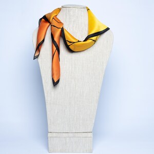 K&ouml;nnte beinhalten: Ein Seidenschal in Orange und Gelb mit schwarzen Verzierungen, drapiert auf einer Schaufensterpuppe. Der Schal hat ein geometrisches Design und ist am Hals geknotet, wobei die Enden herunterh&auml;ngen.