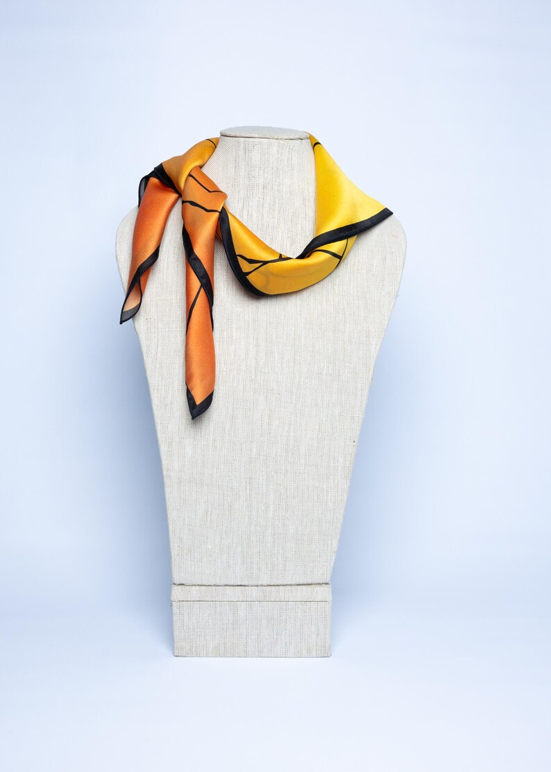 K&ouml;nnte beinhalten: Ein Seidenschal in Orange und Gelb mit schwarzen Verzierungen, drapiert auf einer Schaufensterpuppe. Der Schal hat ein geometrisches Design und ist am Hals geknotet, wobei die Enden herunterh&auml;ngen.