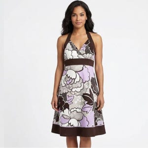 Puede incluir: Vestido con cuello halter con estampado floral en tonos marrón, blanco y lavanda. El vestido tiene cuello en V, cinturilla marrón y bajo marrón. El vestido, a la altura de la rodilla, tiene una falda acampanada.