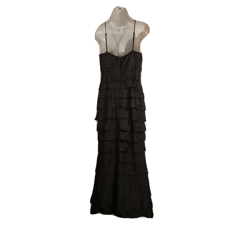 May include: A dark brown, floor-length dress with a tiered, ruffled design. The dress features thin spaghetti straps and a fitted bodice. The back of the dress is visible, showcasing the zipper closure. The dress is displayed on a mannequin.