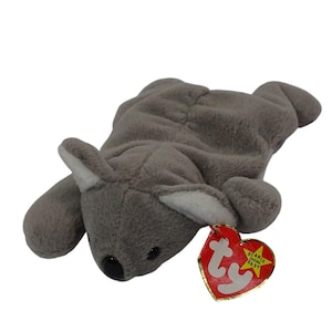 May include: A gray plush koala toy with white inner ears and a black nose. The toy has a red heart-shaped tag with gold trim and the words "Beanie Baby" printed on it. The koala is lying down.
