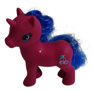 May include: A pink toy pony with a blue mane and tail. The pony has a horn and a cherry graphic on its side. The eyes are detailed with eyelashes and a star design. The toy is standing on all four legs.